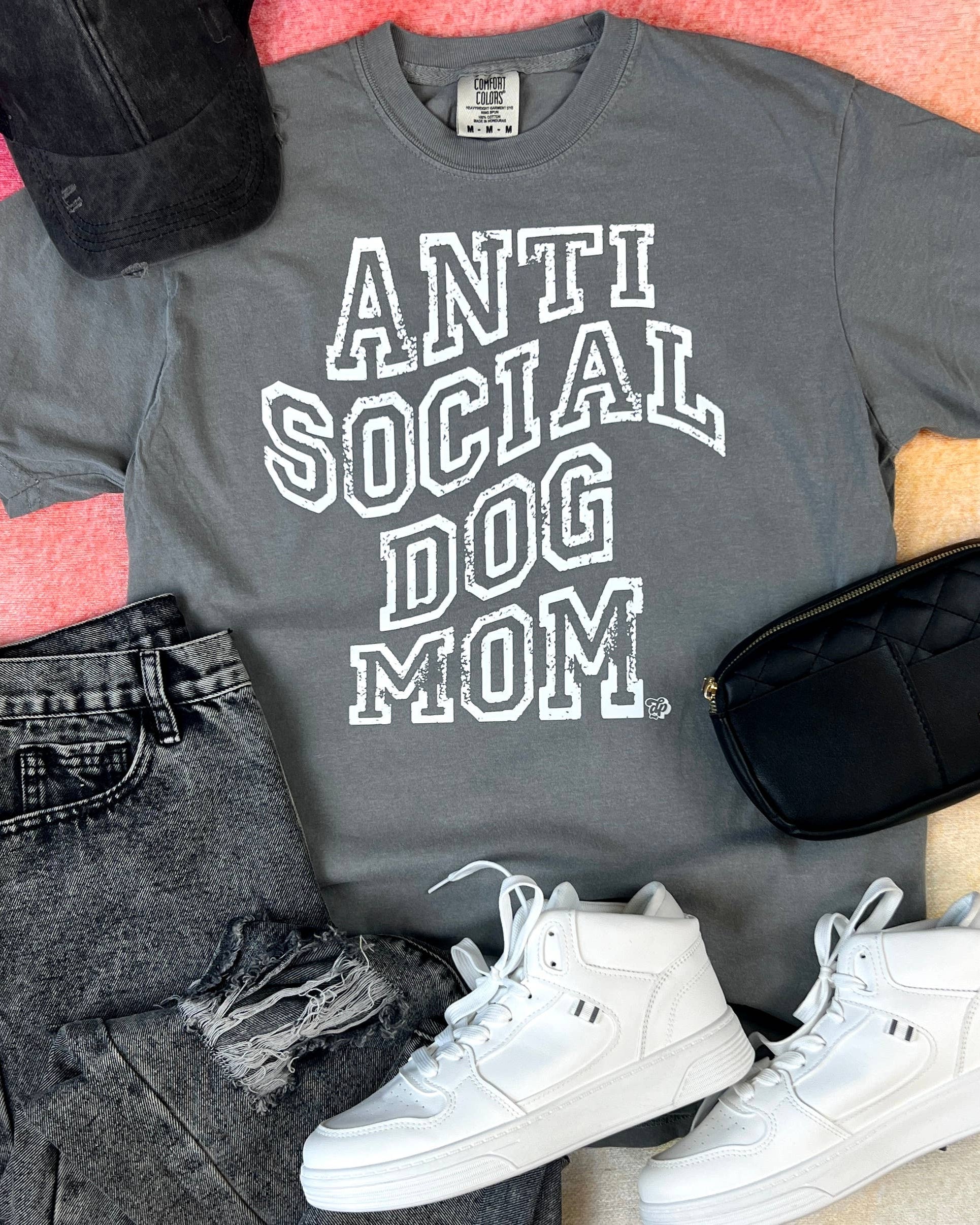 Gray t-shirt with 'ANTI SOCIAL DOG MOM' text, black jeans, white sneakers, and a black bag on a pink surface.
