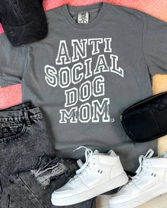 Gray t-shirt with 'ANTI SOCIAL DOG MOM' text, black jeans, white sneakers, and a black bag on a pink surface.